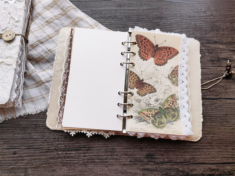 May include: A brown leather bound journal with white lace trim and a page featuring a vintage illustration of butterflies. The journal has a ring binder and a blank page.