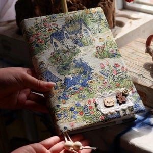 Könnte beinhalten: Ein stoffbezogenes Notizbuch mit einer idyllischen Szene mit Häusern, Blumen und Bäumen in Blau-, Grün- und Rosatönen. Das Notizbuch hat eine Spitzen- und Holzverzierung mit einem Katzenkopf und einem Bären. Eine Quaste mit Holzperlen hängt am Notizbuch.