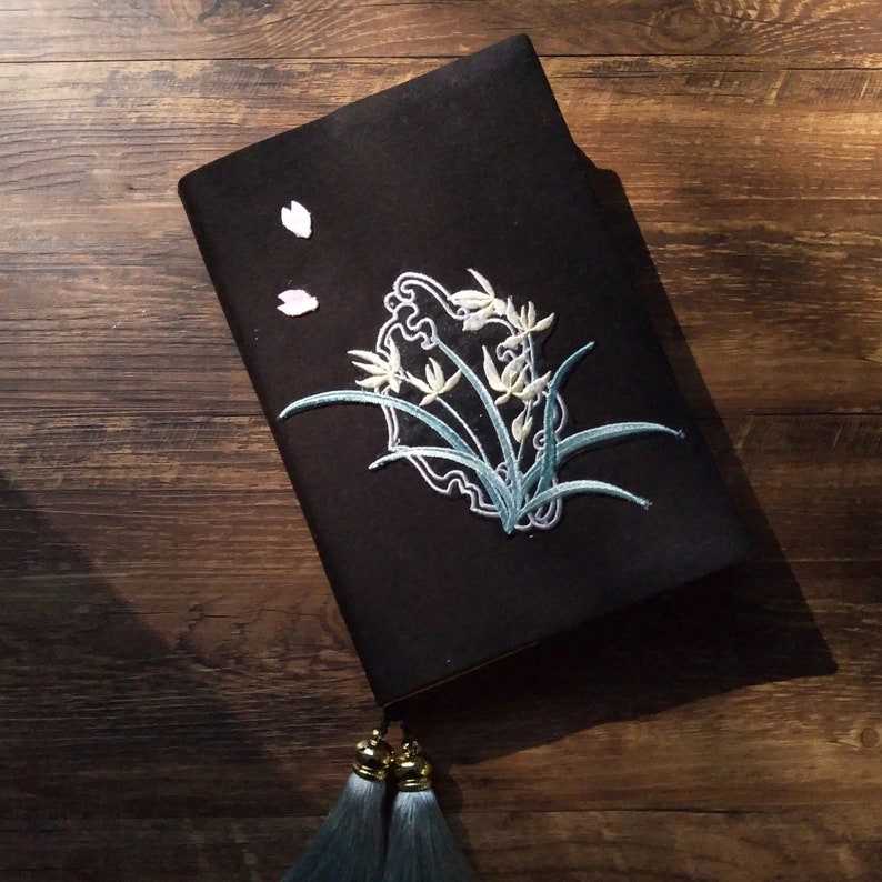 May include: A black notebook with a floral embroidery design. The notebook has a tassel and is closed. The notebook is on a wooden surface.