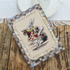 May include: A rectangular fabric-covered journal featuring a blue, brown, and white chequered pattern. The centre displays a beige fabric patch with a lace border, showcasing a printed illustration of a rabbit wearing a heart-patterned vest.