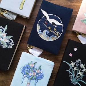 May include: A collection of six black and blue fabric journals with embroidered designs. The designs include a crane, flowers, and a geometric pattern. The journals have tassels and are tied with ribbon.