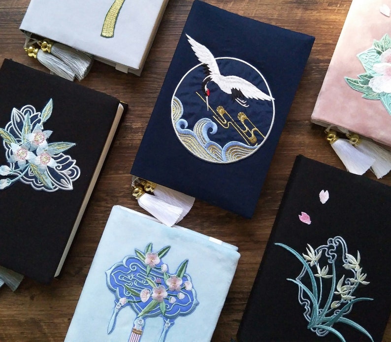 May include: A collection of six black and blue fabric journals with embroidered designs. The designs include a crane, flowers, and a geometric pattern. The journals have tassels and are tied with ribbon.