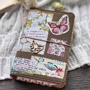 Peut inclure: Journal marron avec un motif de papillon. Le journal présente une couverture en tissu marron avec des patchs décoratifs d'imagerie florale et de papillons. Un charme de papillon en bois et une bande élastique sécurisent le journal. Le texte "A single dream is more powerful than a thousand" est visible.