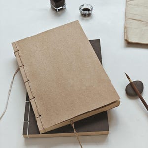 May include: Two handmade journals, one with a brown cover and the other with a dark brown cover, are stacked on top of each other. The journals are bound with a visible stitch and twine. Ink and a brush are also visible.