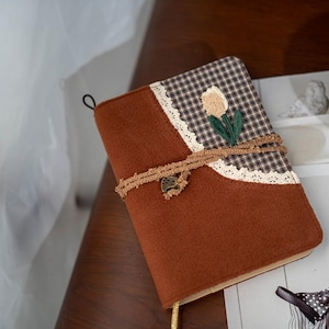 Könnte beinhalten: Ein braunes Notizbuch mit floralem Design. Das Cover hat ein braunes Wildleder-ähnliches Material, ein Karomuster und eine cremefarbene Tulpenstickerei. Ein dekoratives Seil umschlingt das Notizbuch und ist mit einem Charm befestigt.