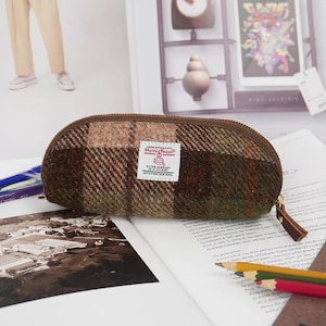 May include: A brown and green plaid pencil case made of Harris Tweed wool fabric. The case features a brown zipper closure with a matching leather pull tab and a white rectangular label on the front that reads Harris Tweed Outer Hebrides.
