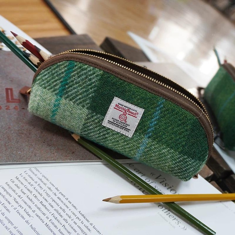 May include: A semi-circular pencil case made of green Harris Tweed wool fabric with a plaid pattern, featuring a brown zipper closure and a white rectangular label that reads Harris Tweed Outer Hebrides of Scotland 100% pure new wool.