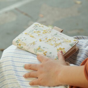 May include: A small, white notebook with a floral embroidery design. The notebook is closed and has a light brown leather cover. The notebook is sitting on a person's lap.