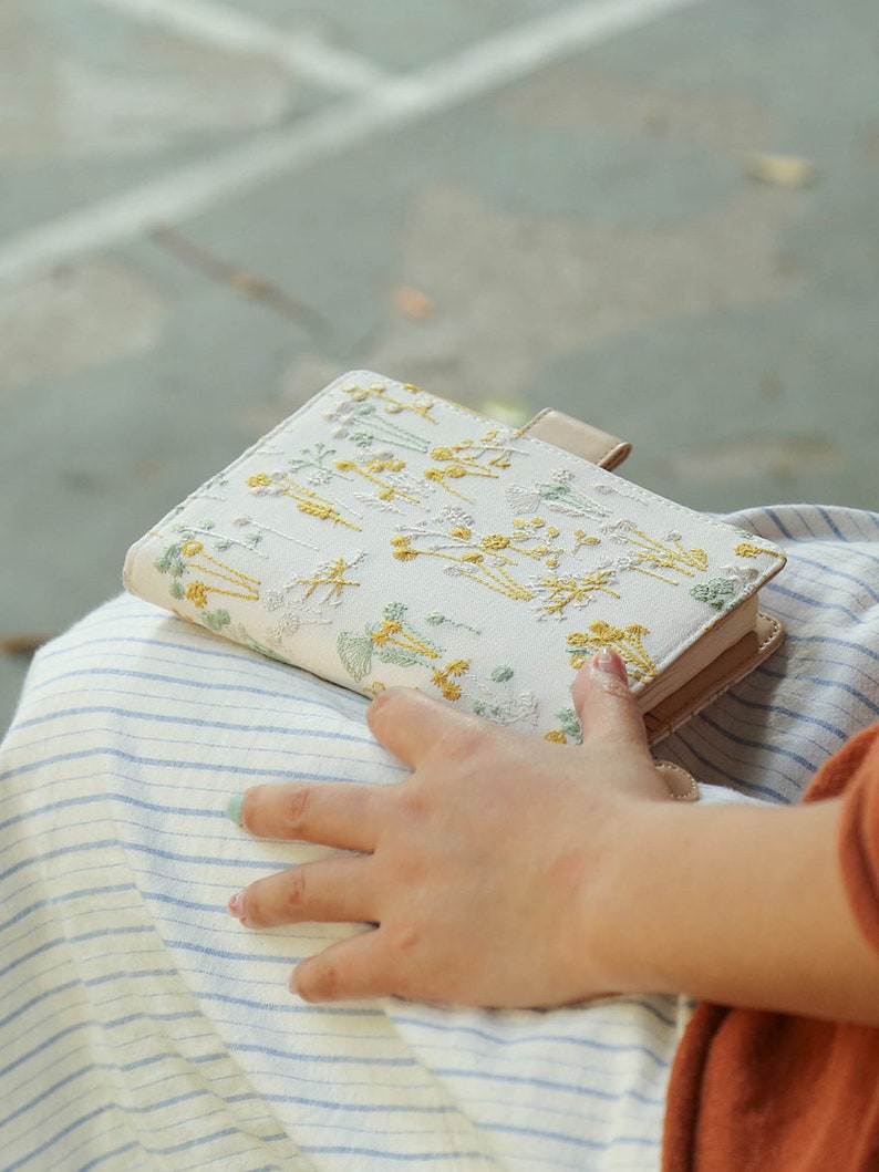 May include: A small, white notebook with a floral embroidery design. The notebook is closed and has a light brown leather cover. The notebook is sitting on a person's lap.