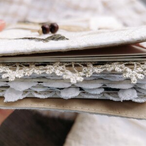May include: A white fabric-covered journal with a decorative lace trim and a brown leather binding. The journal is open to reveal pages with more lace trim.
