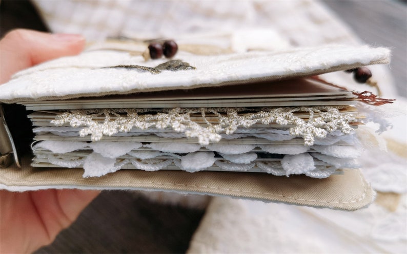 May include: A white fabric-covered journal with a decorative lace trim and a brown leather binding. The journal is open to reveal pages with more lace trim.