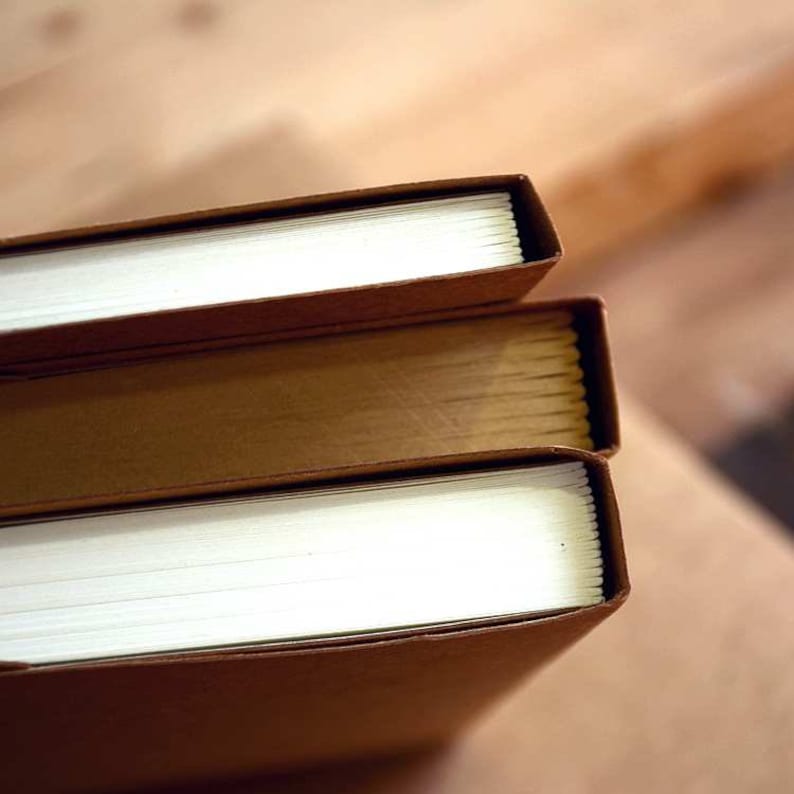 May include: Three brown hardcover books stacked on top of each other. The top book has a white spine with visible pages.