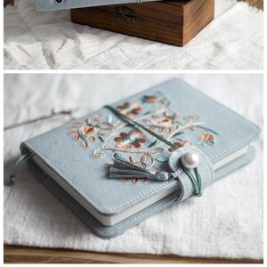 May include: A pale blue journal with orange and brown floral embroidery. It is secured with a green cord and a pearl clasp. The journal is on a wooden box. The text says: 'After the book gradually thickens with use, the knot can be opened to facilitate the book to be closed.'