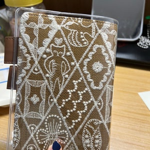 May include: A brown fabric notebook cover with a clear plastic cover. The fabric has a white embroidered pattern of various shapes and designs.