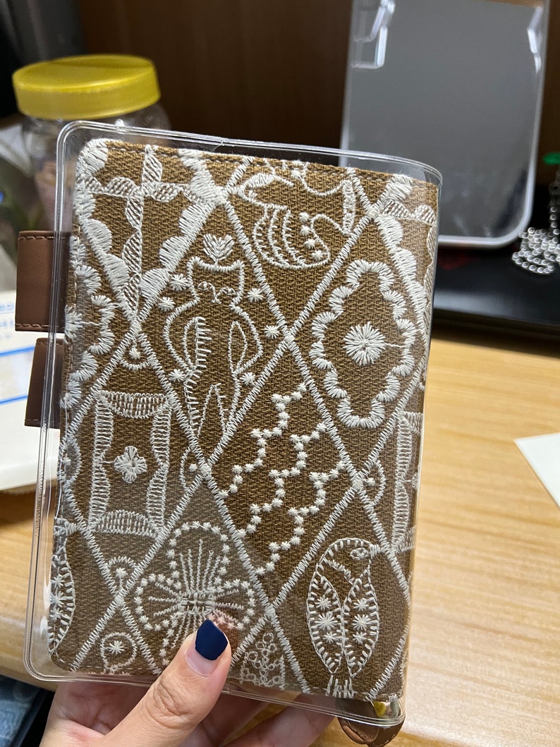 May include: A brown fabric notebook cover with a clear plastic cover. The fabric has a white embroidered pattern of various shapes and designs.