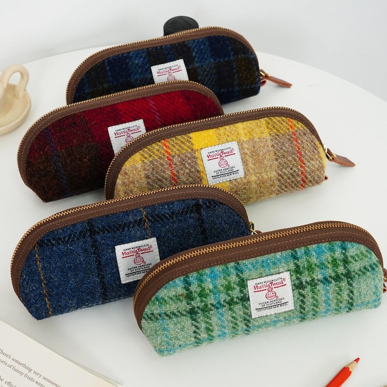 May include: Five semi-circular pencil cases made of Harris Tweed wool fabric in various plaid patterns including blue, red, yellow, dark blue, and green. Each case features a brown zipper, brown trim, and a white Harris Tweed label on the front.