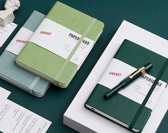 Op de afbeelding: Een verzameling pocket notebooks in groen en blauw, met een pen erop. Elk notitieboekje heeft een witte band met het woord "POCKET" en "PAPER IDEAS" erop gedrukt. De notebooks liggen op een wit oppervlak.