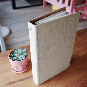 Pode incluir: Um fichário de linho bege com um mecanismo de argolas e um inserto de papel branco. O fichário está sobre uma superfície de madeira.