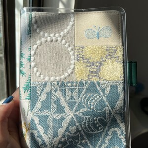 May include: A clear plastic cover with a fabric insert. The fabric is a patchwork of different patterns, including a butterfly, a floral design, and a geometric pattern. The fabric is in shades of white, blue, and green.