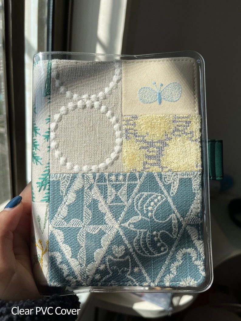 May include: A clear plastic cover with a fabric insert. The fabric is a patchwork of different patterns, including a butterfly, a floral design, and a geometric pattern. The fabric is in shades of white, blue, and green.