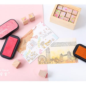 May include: A set of 12 wooden stamps with floral designs, two black ink pads, and a card with a London theme. The stamps are in a wooden box with a lid. The ink pads are rectangular and have a red and orange ink pad.