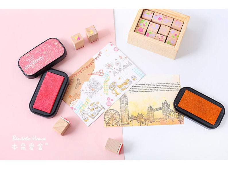 May include: A set of 12 wooden stamps with floral designs, two black ink pads, and a card with a London theme. The stamps are in a wooden box with a lid. The ink pads are rectangular and have a red and orange ink pad.