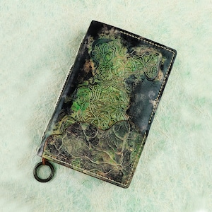 Könnte beinhalten: Ein schwarzes Lederjournal mit einem geprägten floralen Design in Grün und Braun. Das Journal hat einen Metallring, der an der Bindung befestigt ist.