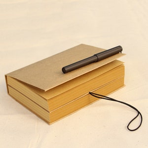 May include: A brown notebook with a black pen on top and a black string attached to the cover. The notebook is closed and has a textured cover.