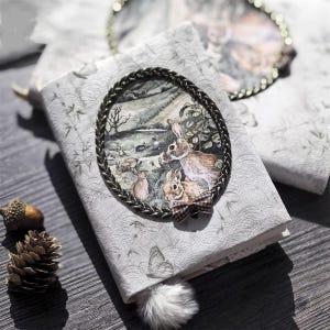 Puede incluir: Un diario decorativo con una cubierta gris claro con un patrón de mariposas repetido. Una imagen ovalada de conejos en una escena forestal está enmarcada con un borde trenzado. Un pequeño lazo decorativo y un pompón blanco y esponjoso añaden detalles.