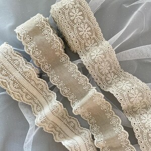 May include: Three beige cotton lace trims with delicate floral embroidery. The lace trims are arranged on a white tulle background.