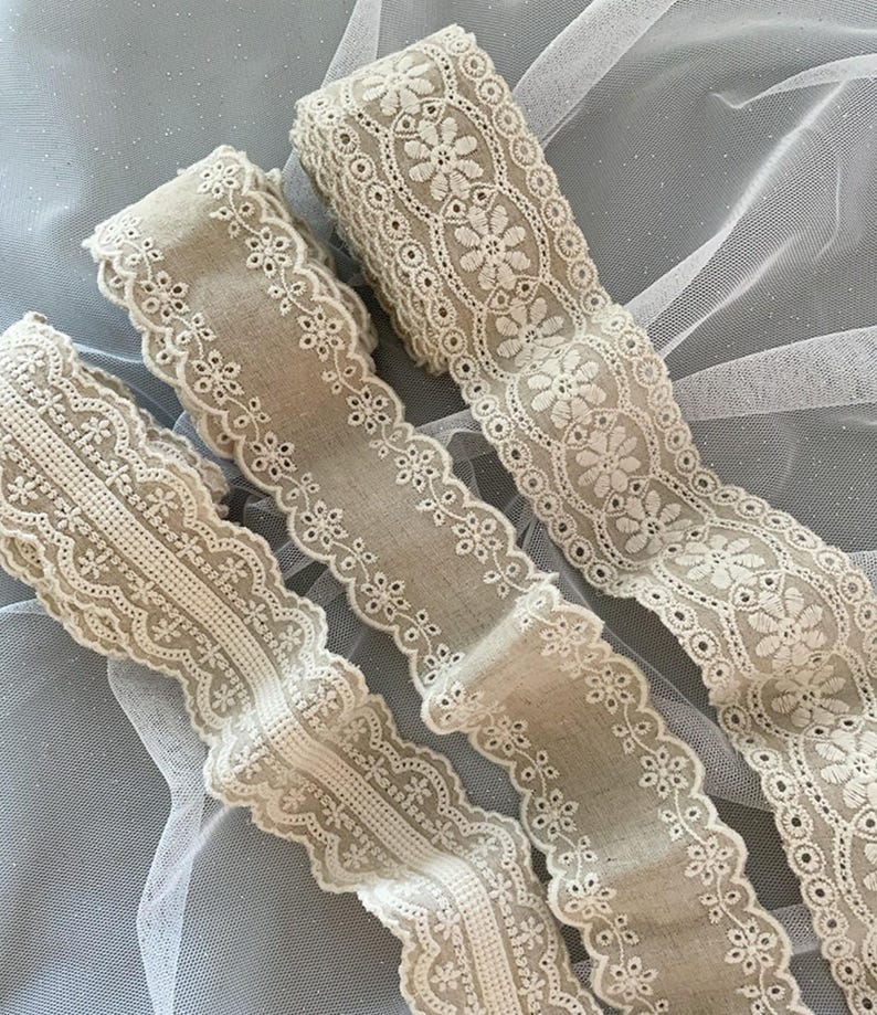 May include: Three beige cotton lace trims with delicate floral embroidery. The lace trims are arranged on a white tulle background.