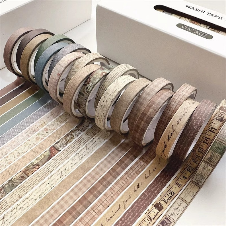 May include: A set of vintage washi tape rolls with various patterns, including floral, plaid, and script designs. The tape is brown, beige, and green. The tape is labeled "Washi Tape Vintage".
