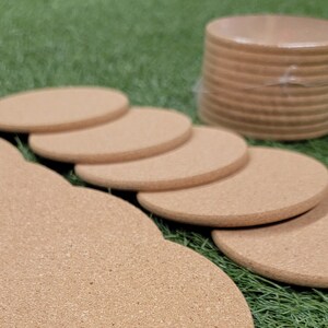 May include: A stack of round cork coasters in various sizes, with the largest coaster in the foreground. The coasters are on a bed of green grass.