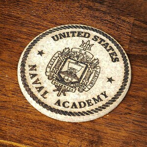 May include: Set of four wooden coasters featuring the United States Naval Academy seal. The seal is a brown and white design with a rope border. The coasters are on a brown wooden surface. (set of 4)