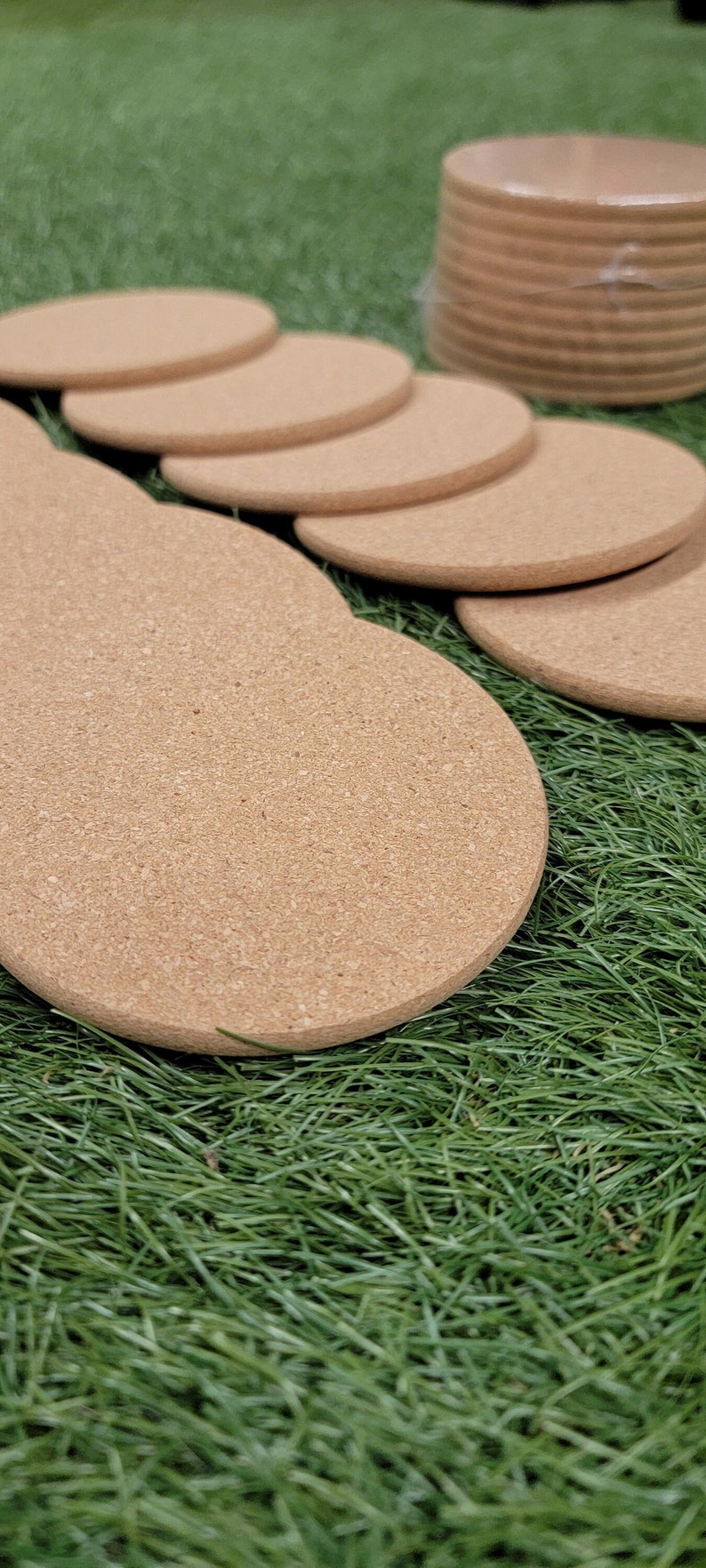 May include: A stack of round cork coasters in various sizes, with the largest coaster in the foreground. The coasters are on a bed of green grass.