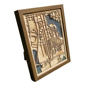 May include: Wooden framed map art featuring a detailed laser-cut city map. The map is made of light-colored wood, showcasing streets, buildings, and waterways. The frame is a dark brown color, and the artwork is displayed at an angle.