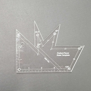 May include: A clear acrylic Perfect Piecer Ruler Template with various angle markings, including 22.5, 30, 45, 60, 120, and 135 degrees. The ruler has inch measurements along one edge and is designed for quilting and fabric cutting.