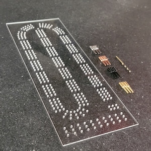 May include: A clear plastic sheet with rows of small holes, used for creating custom jewelry. The sheet is accompanied by a variety of small metal pins in different colors, including gold, silver, and black.