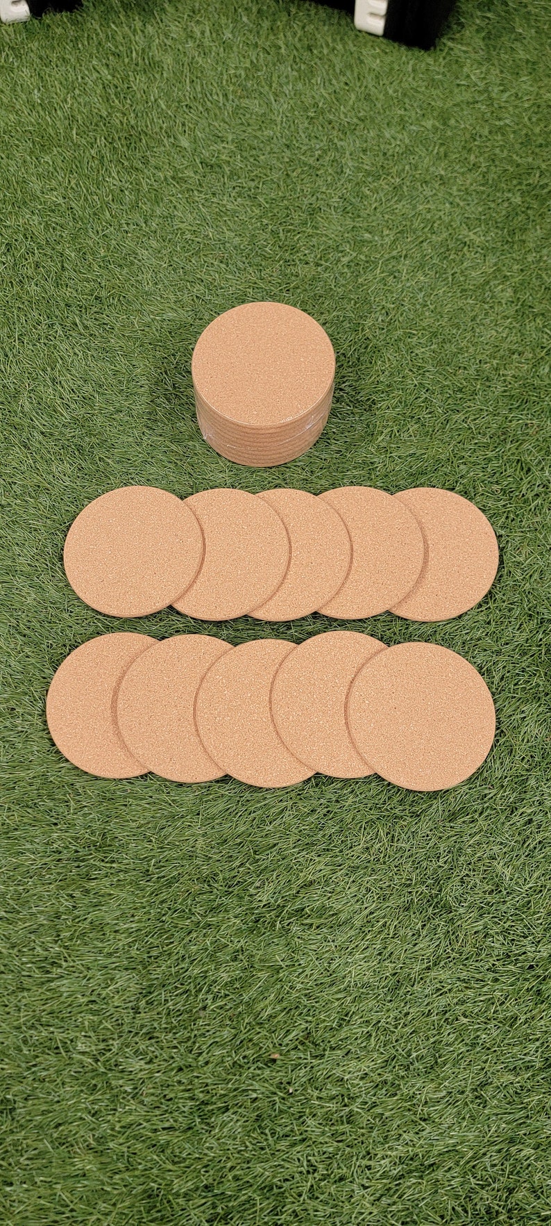 May include: A stack of 12 round cork coasters in a light brown color. The coasters are arranged in a circle on a green grassy surface.