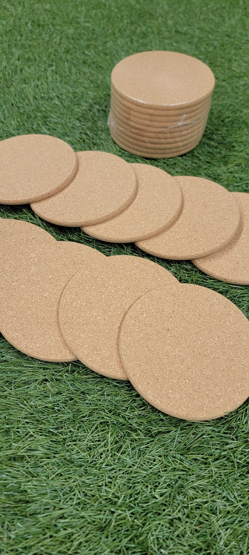 May include: A set of 10 round cork coasters in a natural brown color. The coasters are arranged in a circle on a green artificial grass surface.