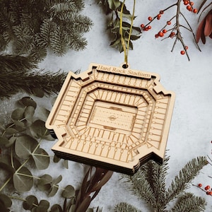 May include: A wooden ornament shaped like Hard Rock Stadium, with intricate details of the field and seating. The stadium name is engraved at the top. The ornament is suspended by a gold string, perfect for holiday decor.