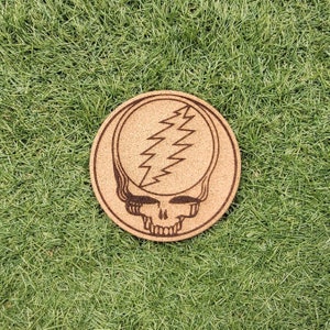 May include: A round cork coaster with a brown wood grain pattern. The coaster features a laser-etched design of a smiling skull with a lightning bolt in the forehead.