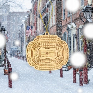 May include: A wooden ornament shaped like the M&T Bank Stadium, home of the Baltimore Ravens. The ornament is brown and features the stadium's seating chart with the team's logo in the center.