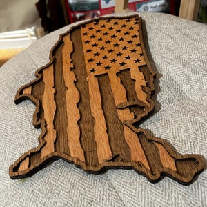 K&ouml;nnte beinhalten: Ein Holzausschnitt der Vereinigten Staaten von Amerika in Form einer Karte, mit dem Design der amerikanischen Flagge in die Karte integriert. Das Holz ist dunkelbraun mit einer helleren braunen Holzmaserung.