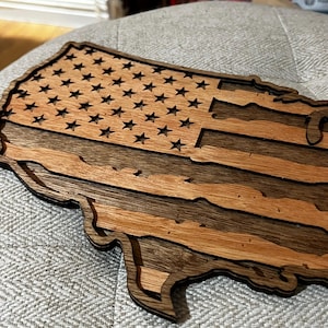K&ouml;nnte beinhalten: Ein Holzausschnitt der Vereinigten Staaten von Amerika in Form einer Flagge. Die Sterne und Streifen sind aus dem Holz geschnitten, wodurch ein dreidimensionaler Effekt entsteht.