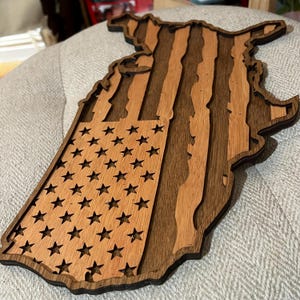 K&ouml;nnte beinhalten: Ein Holzausschnitt der Vereinigten Staaten in Form einer Karte mit dem Design der amerikanischen Flagge. Die Flagge besteht aus zwei verschiedenen Holzarten, einer hellen und einer dunklen, um die Streifen und Sterne zu erzeugen.