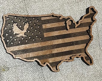 May include: A wooden cutout of the United States of America with the American flag design. The flag is made of dark and light wood, creating a striped pattern. A silhouette of an eagle is in the center of the stars.