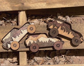 May include: Three wooden toy trucks with mountain and forest designs. The trucks are made of layered wood in shades of brown and tan, with intricate wheel designs. The trucks are arranged on a wooden surface with leaves.