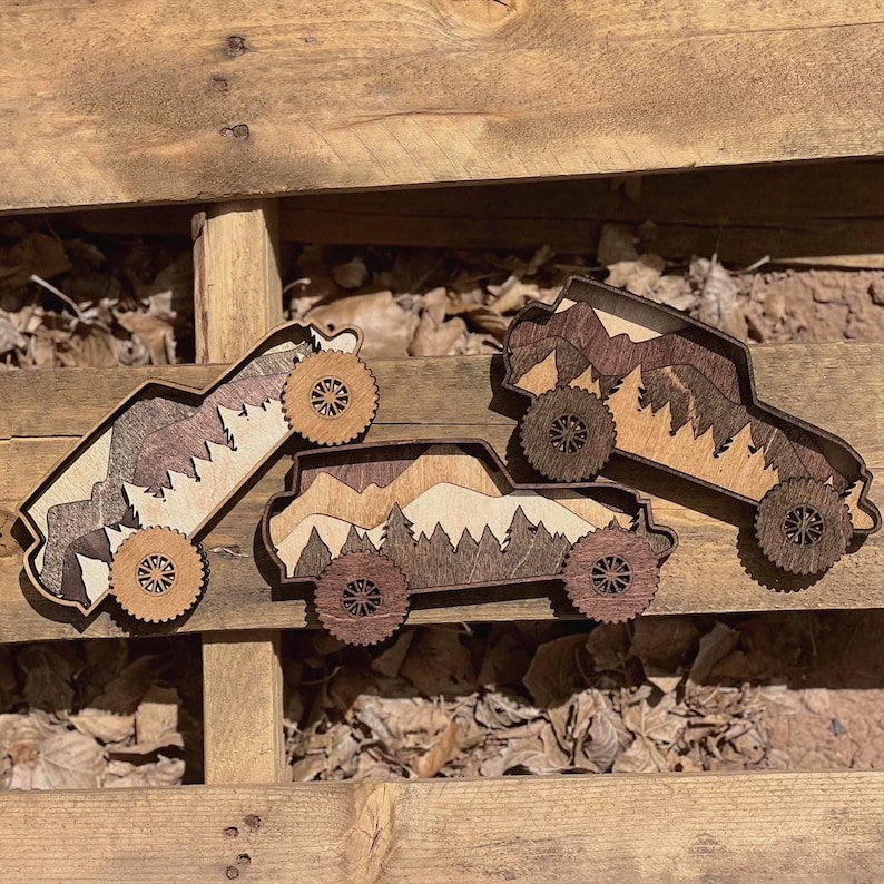 May include: Three wooden toy trucks with mountain and forest designs. The trucks are made of layered wood in shades of brown and tan, with intricate wheel designs. The trucks are arranged on a wooden surface with leaves.