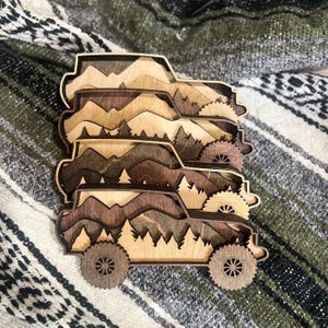 May include: Stacked wooden cutouts of off-road vehicles with mountain and forest scenes. The layered design features varying shades of brown and tan, with detailed wheels and tire treads. The overall theme is nature and adventure.